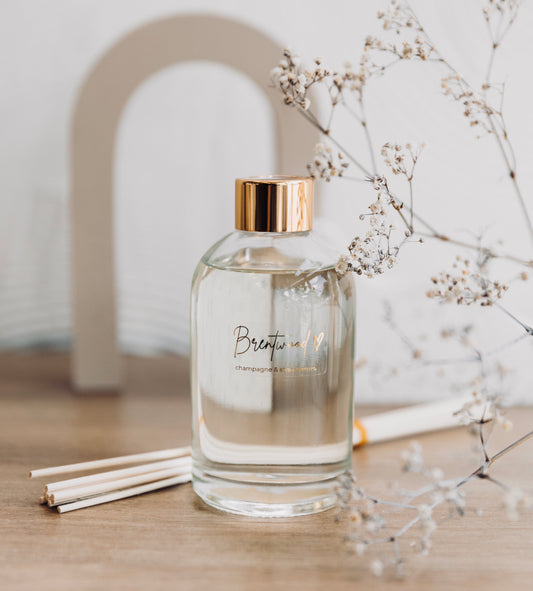 A reed diffuser in a glass bottle and shiny gold cap, filled with fragrant liquid and decorated with a bundle of thin reeds. The diffuser is placed on a wooden surface, with dried flowers in the foreground, creating a serene, whimsical and elegant display.
