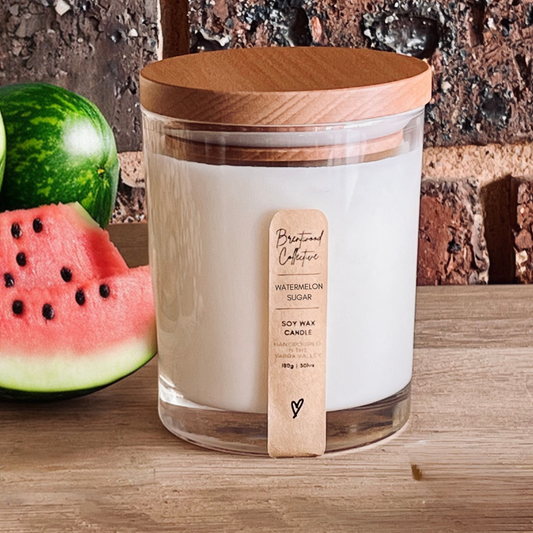 Watermelon Sugar scented soy wax candle. Candle is in clear jar with timber lid, sitting on an aesthetically pleasing wooden bench top with a dark brick wall in the background. Also with a juicy watermelon sitting on the left side behind the candle