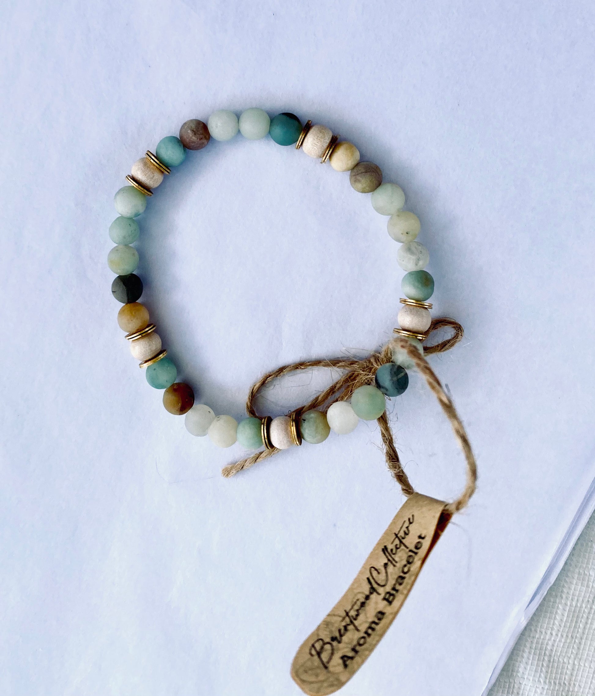 Single amazonite diffuser bracelet made from 6mm beads, with brass heishi disc embellishments and natural wood beads. Laid on white background with Kraft label tag.