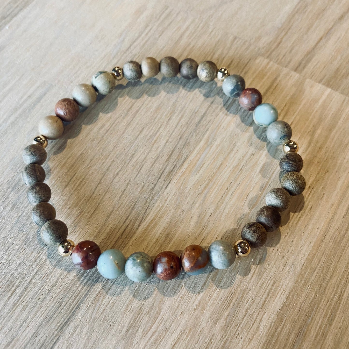 Aqua Terra Jasper aroma bracelet/diffuser bracelet made from 6mm Aqua Terra Jasper & natural burly wood beads with 18k gold plated embellishments. Laid flat on wood grain surface. 