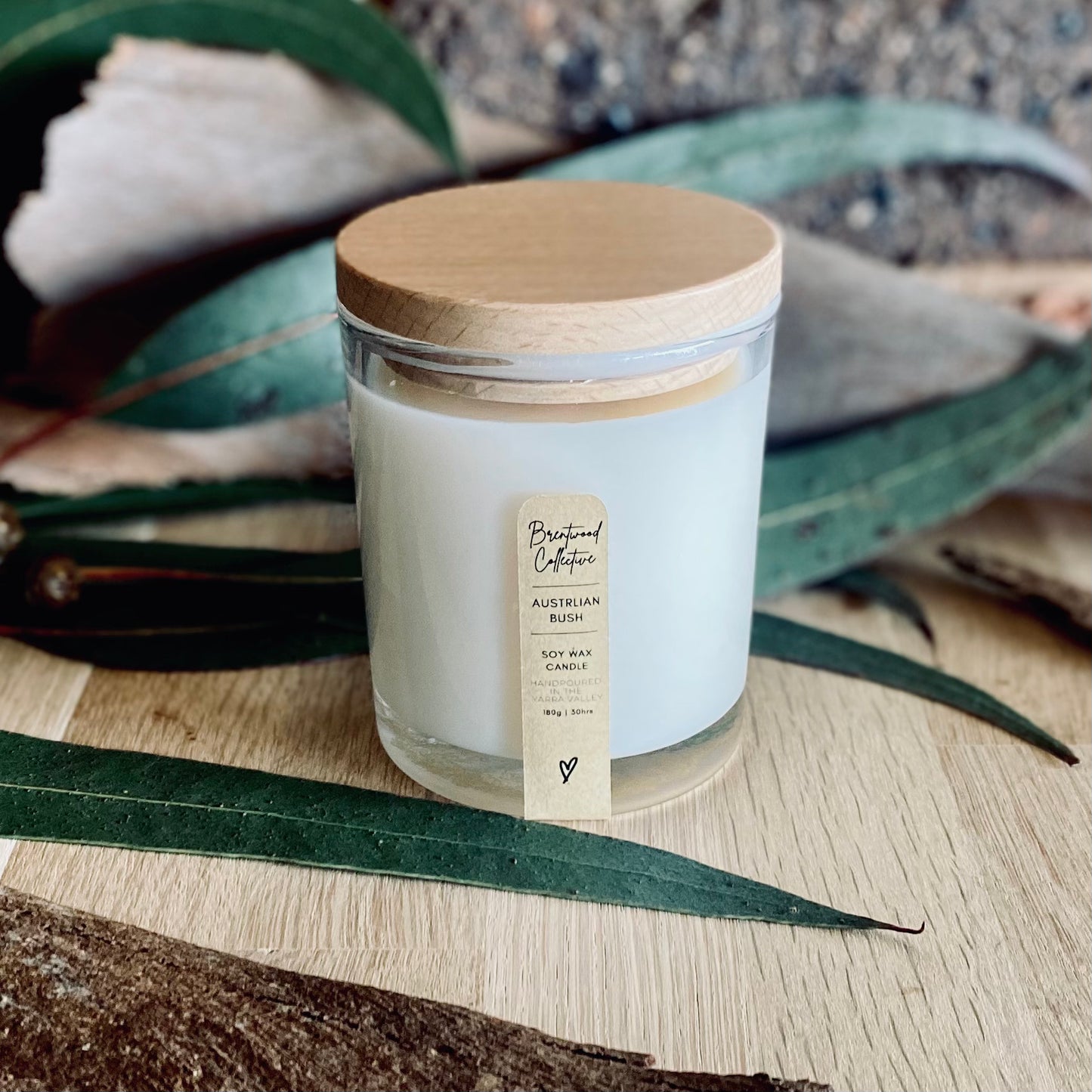 Australian Bush Scented Candle - clear glass jar with natural timber lid & slim/minimal Kraft brown label and black text. Set among Eucalyptus leaves on natural timber bench top with brick wall in background. 