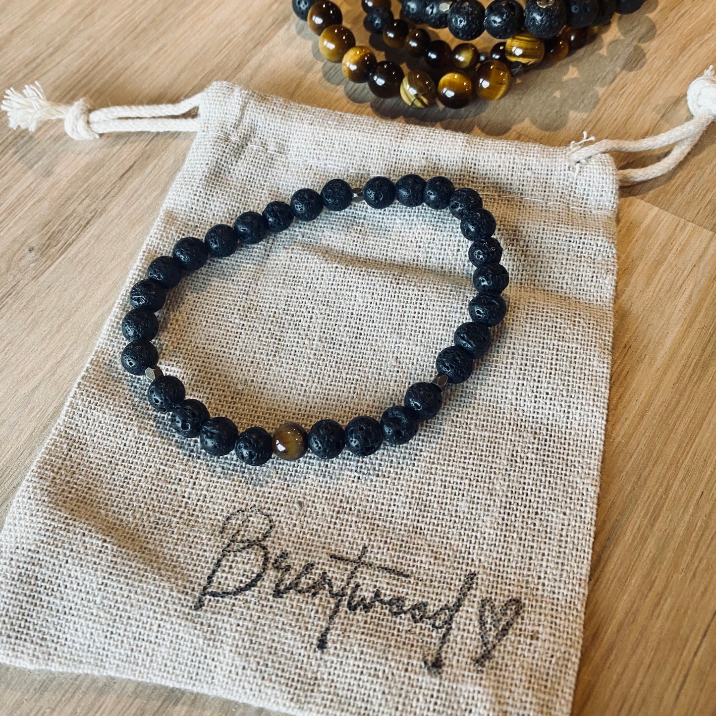 Lava 6 aroma bracelet/diffuser bracelet made from 6mm black lava stone & a single tiger eye bead with antiqued gold faceted embellishments. Laid on cloth pouch/drawstring bag stamped with “Brentwood” and a heart.