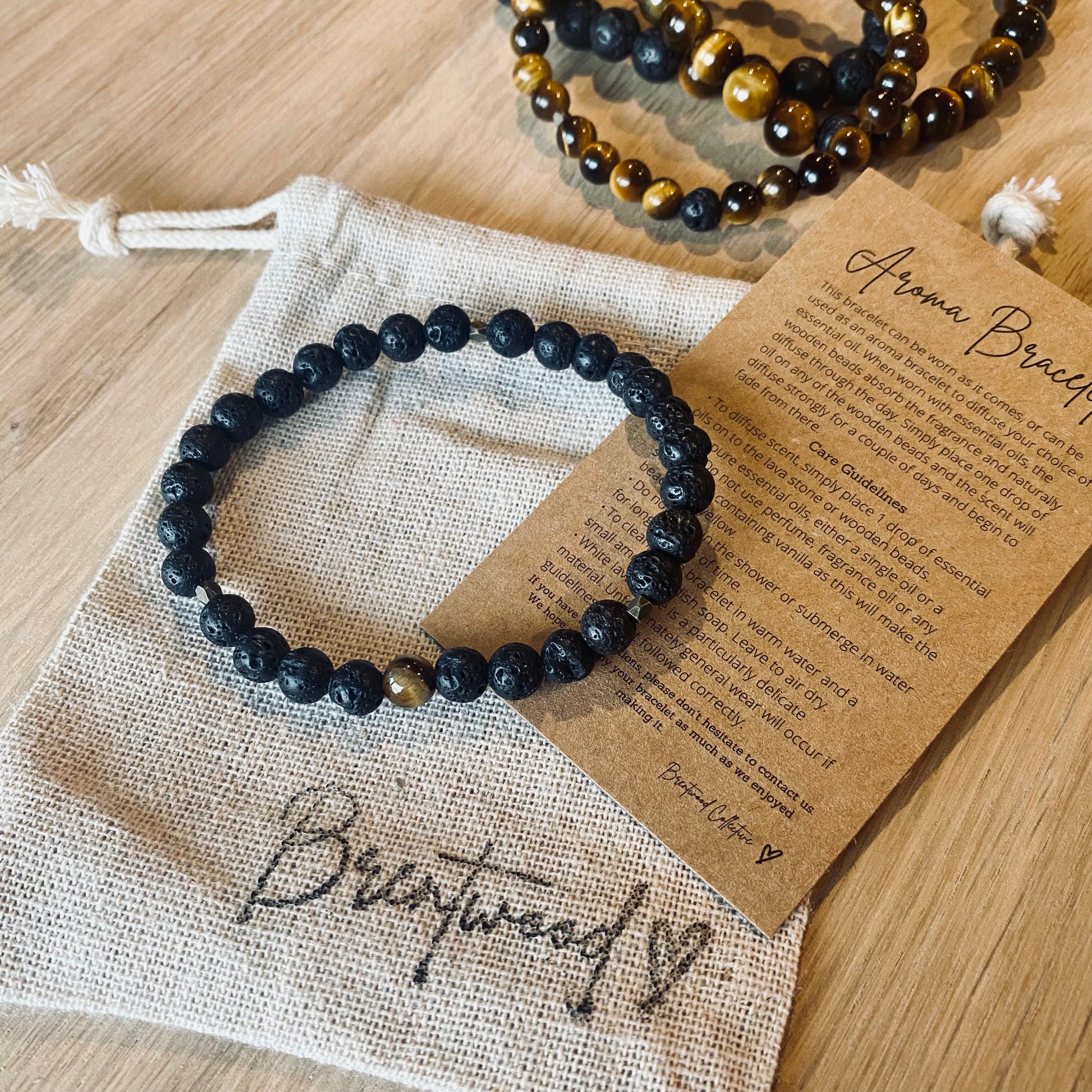 Lava 6 aroma bracelet/diffuser bracelet made from 6mm black lava stone & a single tiger eye bead with antiqued gold faceted embellishments. Laid on cloth pouch/drawstring bag stamped with “Brentwood” and an aroma bracelet information and care card on kraft brown card.