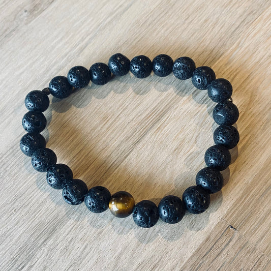 Lava 8 Aroma Bracelet - 8mm beaded diffuser bracelet made from black lava stone & a single tiger eye bead with antiqued gold faceted embellishments. Laid flat on wood grain surface. 