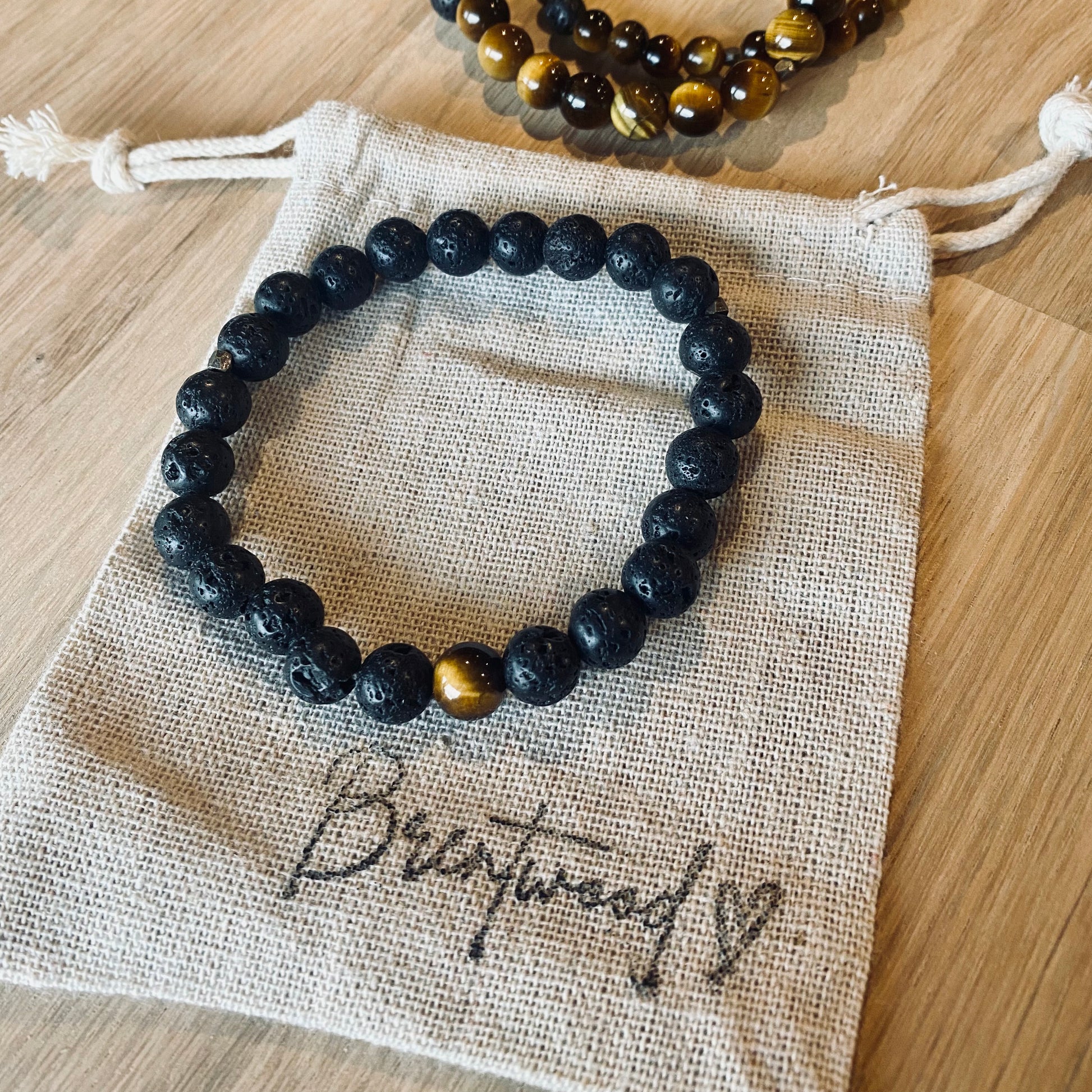 Lava 8 Aroma Bracelet - 8mm beaded diffuser bracelet made from black lava stone & a single tiger eye bead with antiqued gold faceted embellishments. Laid on cloth pouch/drawstring bag stamped with “Brentwood” and a heart.