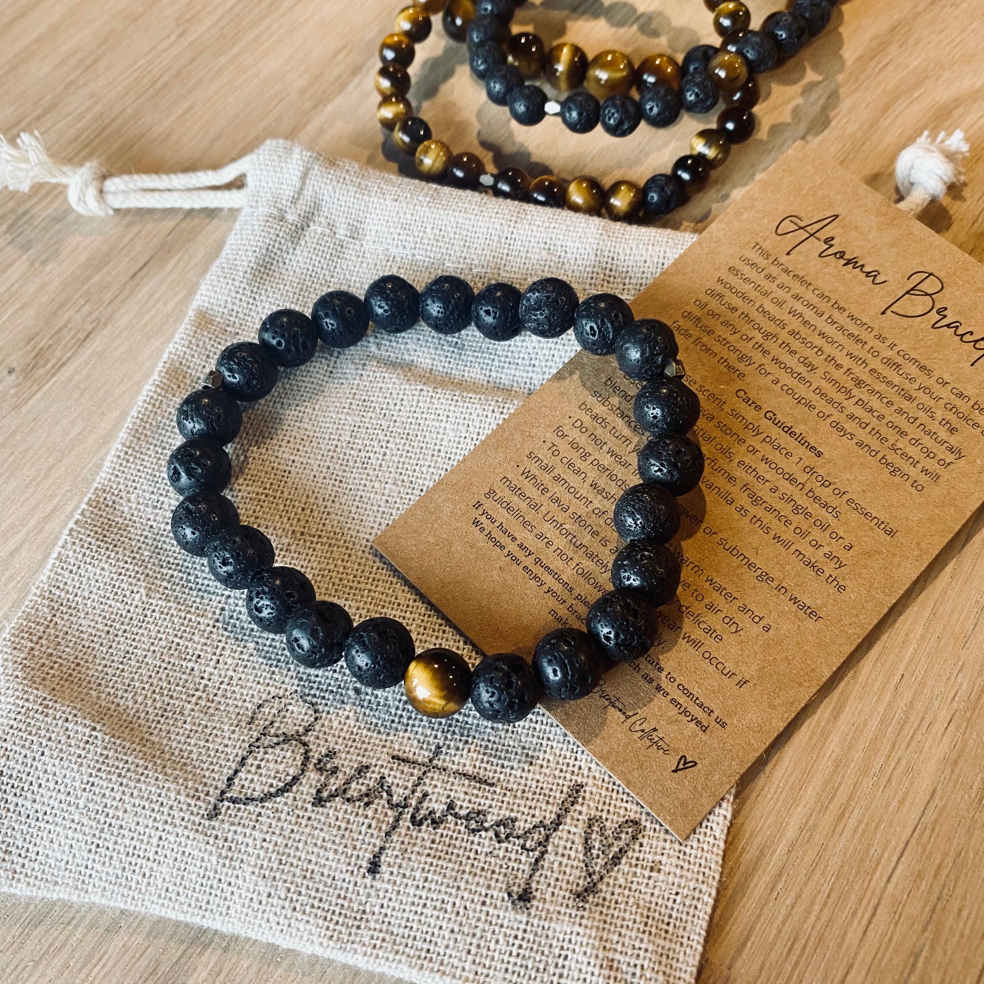 Lava 8 Aroma Bracelet - 8mm beaded diffuser bracelet made from black lava stone & a single tiger eye bead with antiqued gold faceted embellishments. Laid on cloth pouch/drawstring bag stamped with “Brentwood” and an aroma bracelet information and care card on kraft brown card.