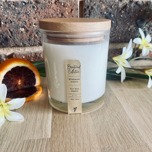 Bohemian Nights Scented Candle - clear glass jar with natural timber lid & slim/minimal Kraft brown label and black text. Set among white & yellow wildflowers and a sliced blood orange. Sitting on a natural timber bench top with brick wall in background. 