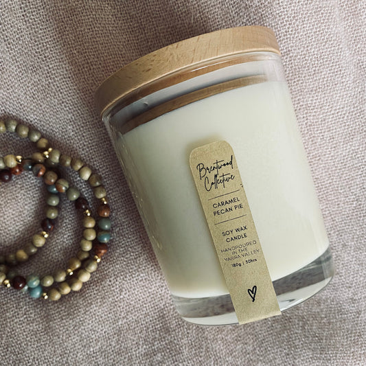 Caramel Pecan Pie Scented Candle - clear glass jar with natural timber lid & slim/minimal Kraft brown label and black text. Laid flat on blush pink fabric background with aqua terra jasper aroma bracelets not the side. 