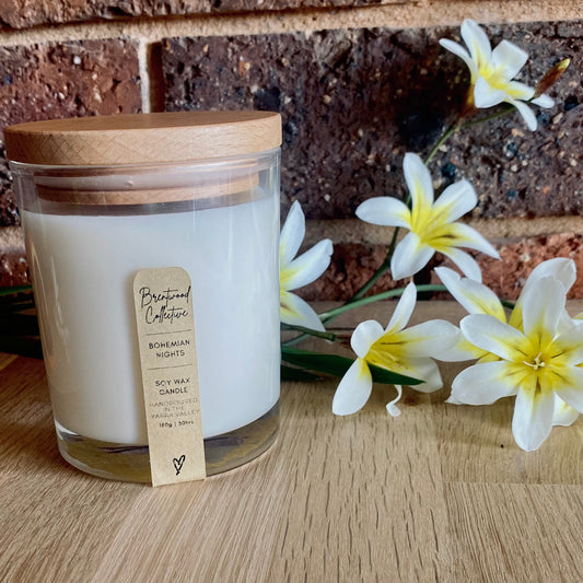 Coconut Lime & Pineapple Scented Candle - clear glass jar with natural timber lid & slim/minimal Kraft brown label and black text. Set among tropical white & yellow flowers on natural timber bench top with brick wall in background. 