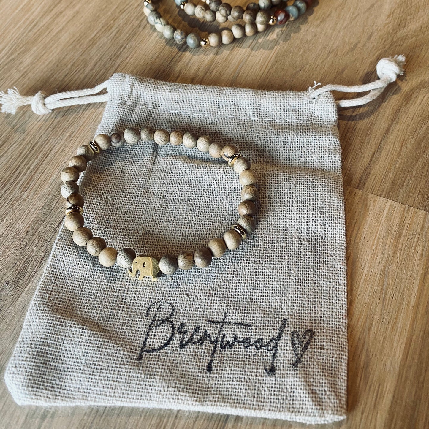 Gold Elephant aroma bracelet/diffuser bracelet made from 6mm burly wood beads beads with gold lucky elephant charm and gold disc embellishments.  Laid on cloth pouch/drawstring bag stamped with “Brentwood” and a heart.