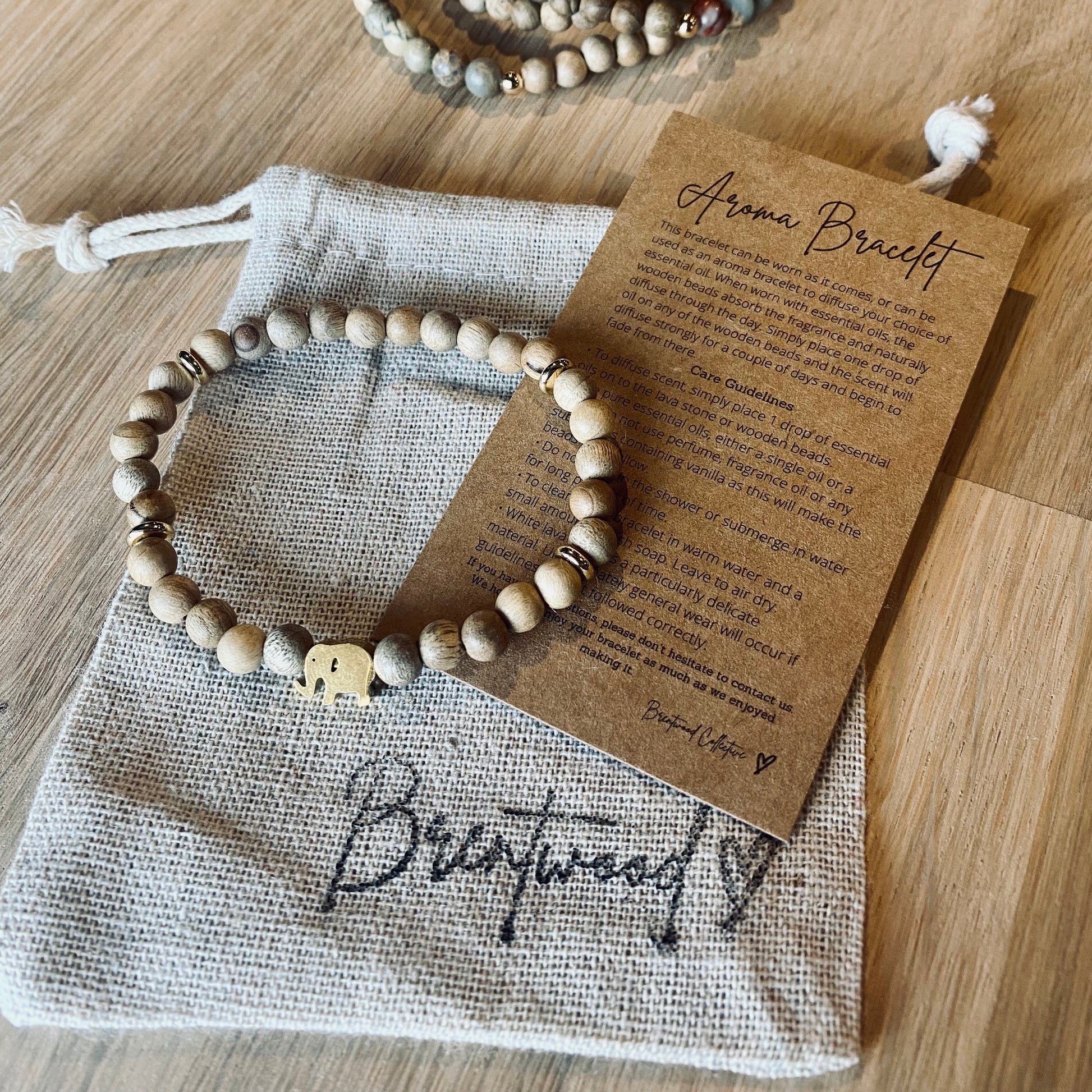 Gold Elephant aroma bracelet/diffuser bracelet made from 6mm burly wood beads beads with gold lucky elephant charm and gold disc embellishments.  Laid on cloth pouch/drawstring bag stamped with “Brentwood” and an aroma bracelet information and care card on kraft brown card.