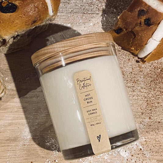 Hot cross bun scented candle in classic size, 180g and 30+ hours burn time. Laid flat on wooden board with hot cross buns, flour and cinnamon.