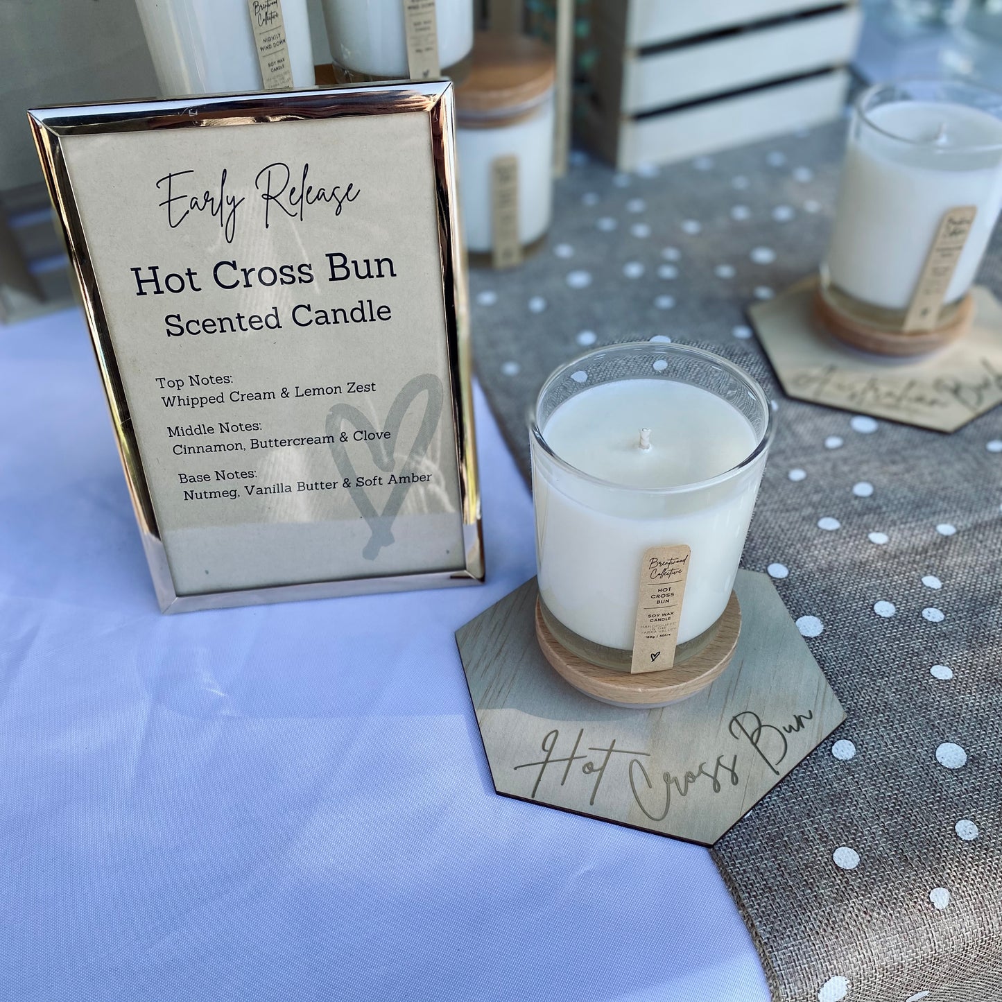 Hot Cross Bun Scented Candle - clear glass jar with natural timber lid & slim/minimal Kraft brown label and black text. Shown on market stall table with hexagon placemat with gold script font reading “Hot Cross Bun”. 