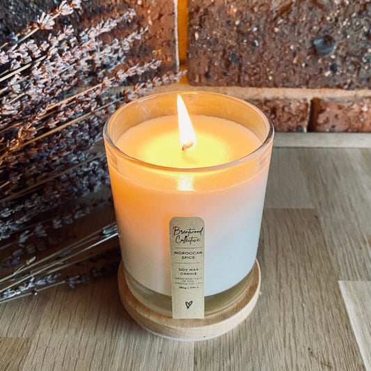 Moroccan Spice Scented Candle - clear glass jar with natural timber lid & slim/minimal Kraft brown label and black text. Candle is burning with large flame. Set among lavender in background on natural timber bench top with brick wall in background. 