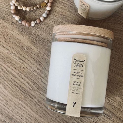 Nightly Wind Down Scented Candle - clear glass jar with natural timber lid & slim/minimal Kraft brown label and black text. Laid flat on natural oak wooden background with mixed aroma bracelets on the side. 
