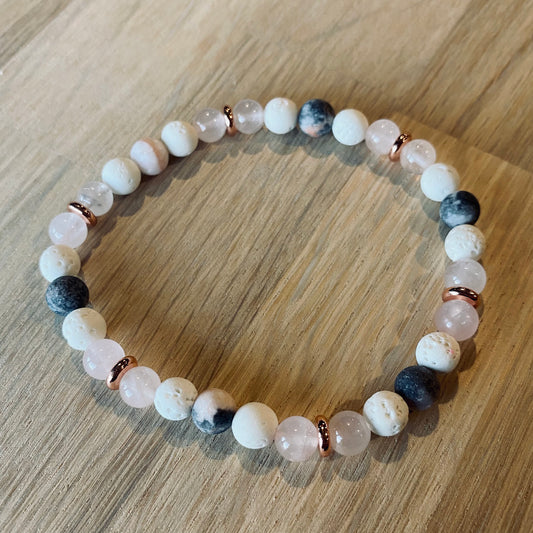 Pink Zebra Jasper Aroma Bracelet - 6mm beaded diffuser bracelet made from a pattern of pink zebra jasper, rose quartz, white lava stone & rose gold disc embellishments. Laid flat on wood grain surface. 