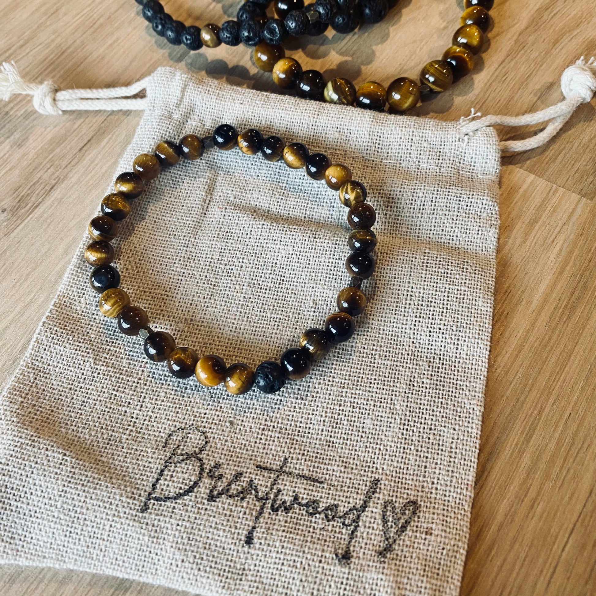 Tiger Eye 6 Aroma Bracelet - 6mm beaded diffuser bracelet made from natural tiger eye gemstone & a single black lava stone bead with antiqued gold faceted embellishments. Laid on cloth pouch/drawstring bag stamped with “Brentwood” and a heart.